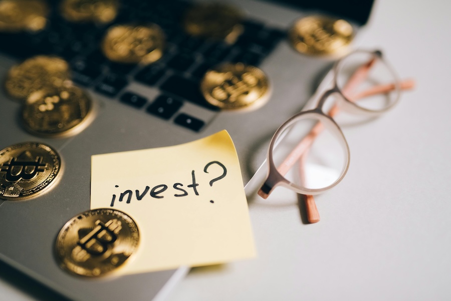 Gold Bitcoin coins scattered on a laptop keyboard next to reading glasses and a yellow sticky note that reads invest?