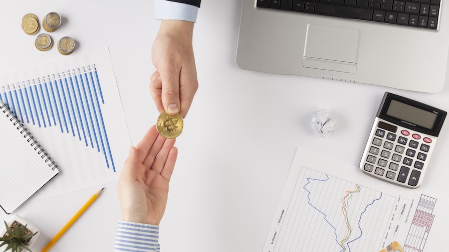 Two Hands Passing A Gold Bitcoin Coin Across A Desk Covered In Printed Market Charts